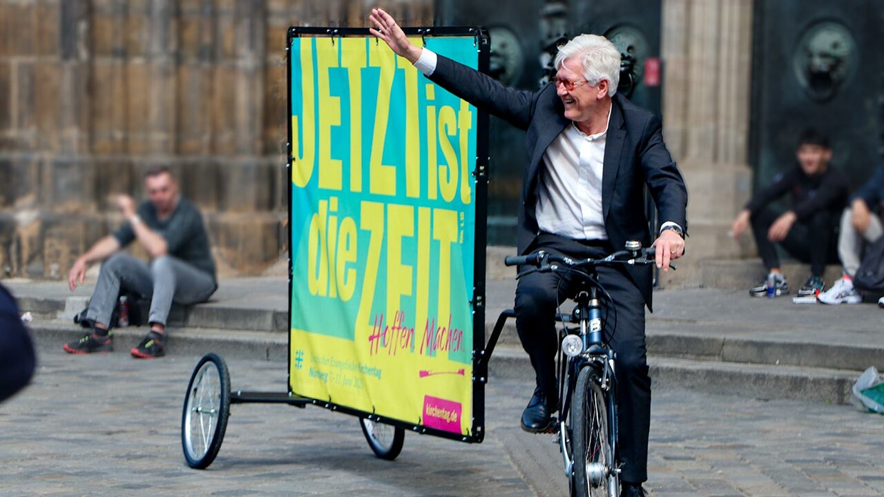 Heinrich Bedford-Strohm auf einem Fahrrad mit Plakat-Anhänger und dem Motiv für den Kirchentag "Jetzt ist die Zeit"