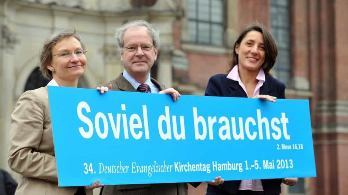 Kirsten Fehrs, Gerhard Robbers und Ellen Ueberschär mit dem großen Schild auf dem "Soviel du brauchst" in weiß auf hellblau steht. Im Hintergrund der Hamburger Michel