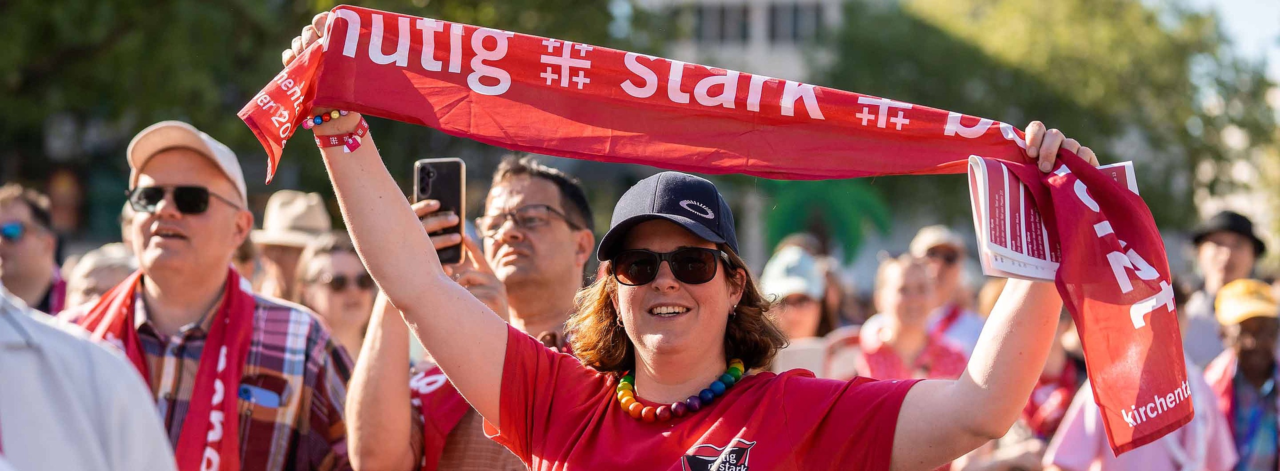 Eine Frau mit Mütze, Sonnenbrille und rotem Kirchentagsshirt steht in der Menschenmenge und hält einen roten Kirchentagsschal in die Luft.