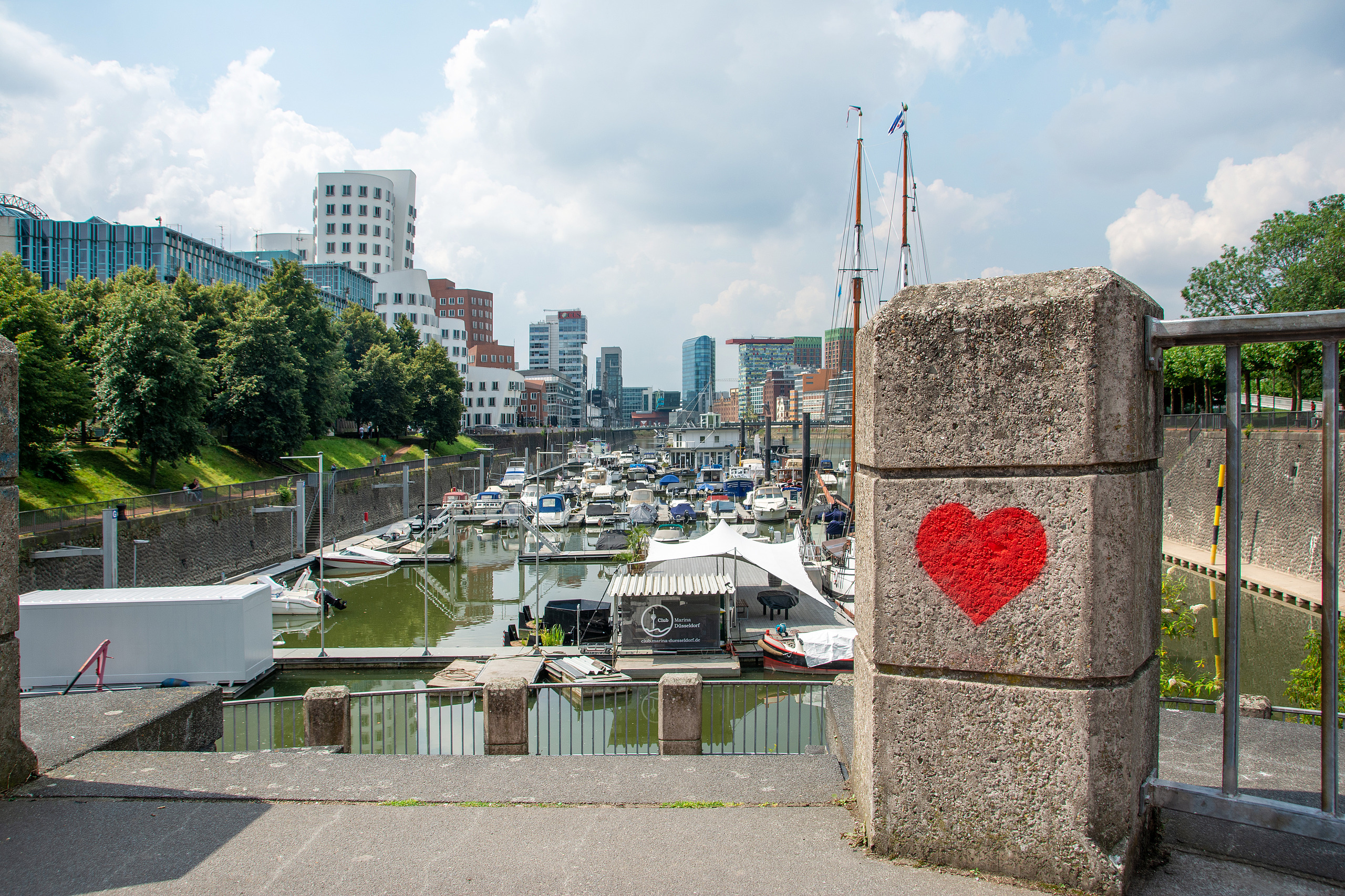 Blick auf den MedienHafen in Düsseldorf mit Hafenbecken, Booten und moderner Architektur. Im Vordergrund ist ein rotes Herz auf einer Steinmauer zu sehen.