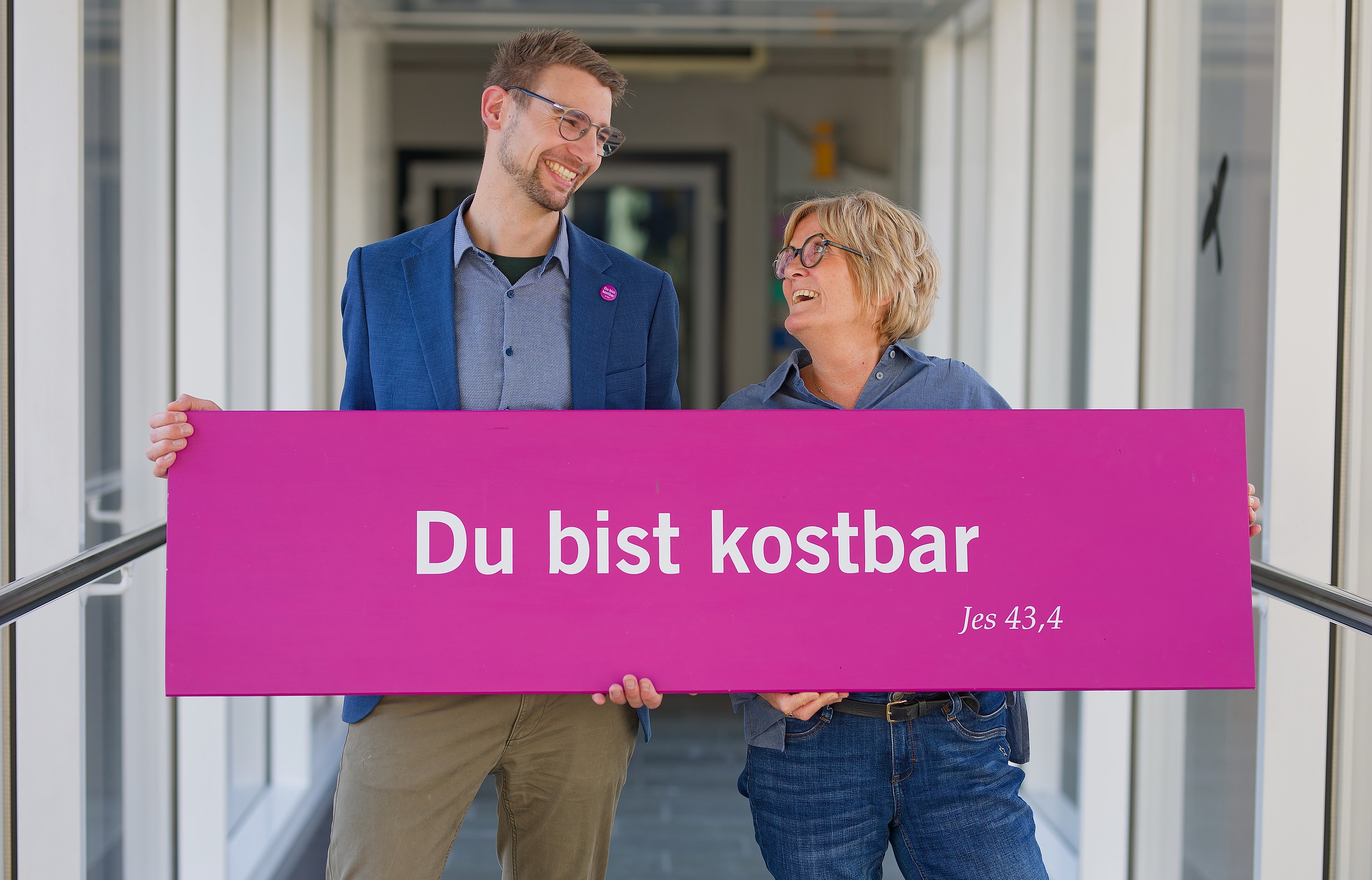 Zwei Personen halten gemeinsam ein pinkes Schild mit der Aufschrift „Du bist kostbar“. Sie stehen in einem hellen Flur und schauen sich lächelnd an.