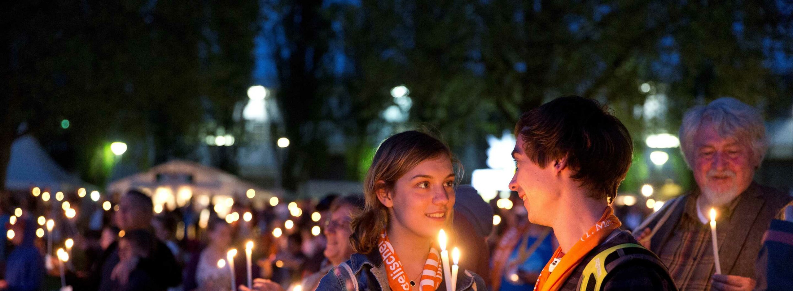 Ein junges Paar schaut sich lächelnd in die Augen während sie die weißen Kerzen zum Abendsegen angezündet zwischen sich halten