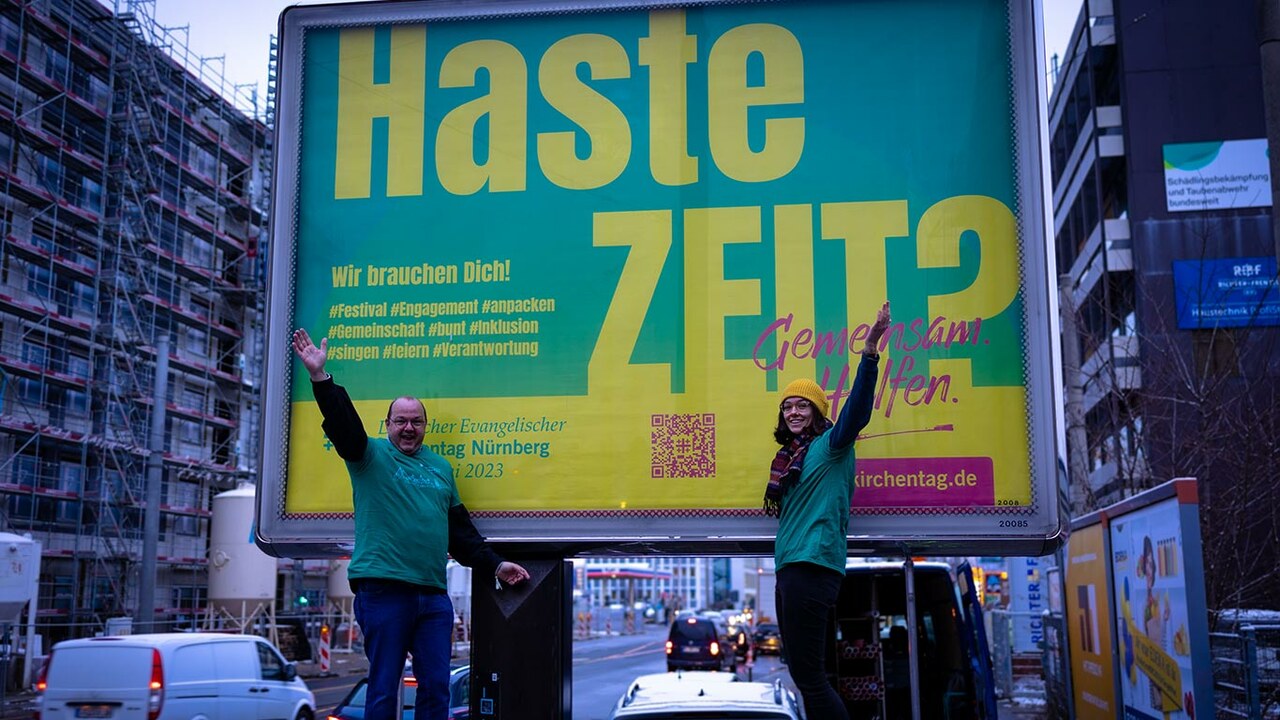 Ein Mitarbeiter und eine Mitarbeiterin des Kirchentages in grünen T-shirts vor einem großen Plakat zum Helfen beim Kirchentag auf einer Straße in Nürnberg.