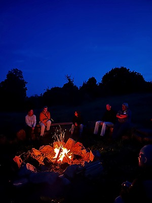 ein Lagerfeuer brennt im Freien, drumherum sitzen Menschen auf Holzbänken