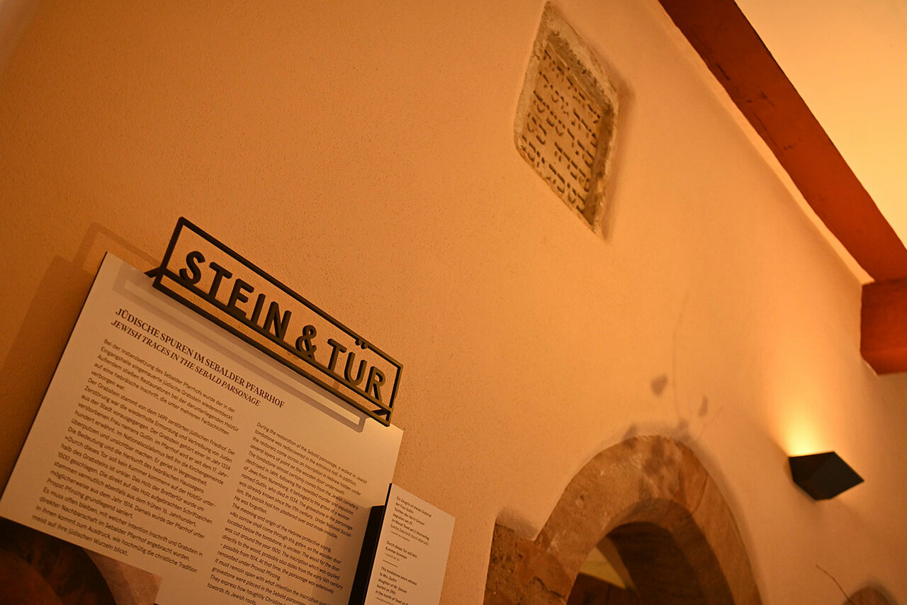 In einer Wand ist oberhalb einer spitz zulaufenden Türe ein Grabstein mit hebräischer Aufschrift zu entdecken. Eine Infografik erklärt die historische Herkunft von Grabstein und Türe.