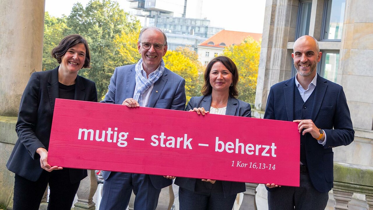 Generalsekretärin Kristin Jahn, Landesbischof der Evangelisch-lutherischen Landeskirche Hannovers Ralf Meister, Kirchentagspräsidentin Anja Siegesmund und Oberbürgermeister der Landeshauptstadt Hannover Belit Onay vor einem rubinroten Schild mit der Losung für Hannover 2025