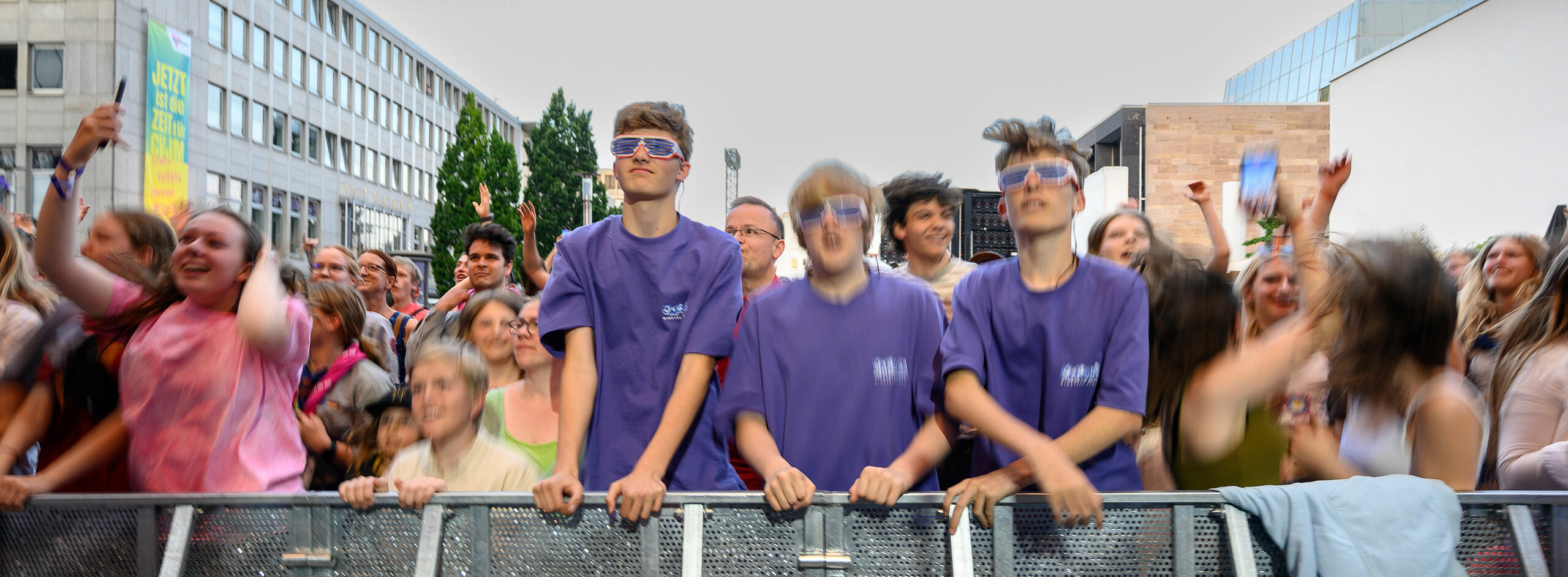 Drei jugendliche Männer in lila T-Shirt stehen in der ersten Reihe eines Konzerts. Der linke trägt eine Sonnenbrille. Die anderen Menschen sind unscharf. Sie tanzen