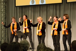 Fünf Frauen singen auf der Bühne in Gebärden, sie sind ganz in schwarz gekleidet und tragen den orangenen Schal des Kirchentages