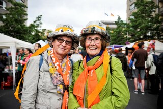 Zwei Damen mit bunten Hüten aus Zeitungen und orangen Schals lachen in die Kamera beim Kirchentag in Berlin 2017 (Abend der Begegnung)