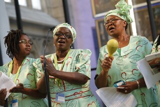 Drei internationale Mitwirkende bei ihrem Auftritt beim Kirchentag, vermutlich afrikanischer Gospel, sie tragen hellgrüne Gewänder, zwei haben passende Kopftücher, eine Rastazöpfe. Die Dame in de Mitte klatscht im Takt, rechts spielt Maracas