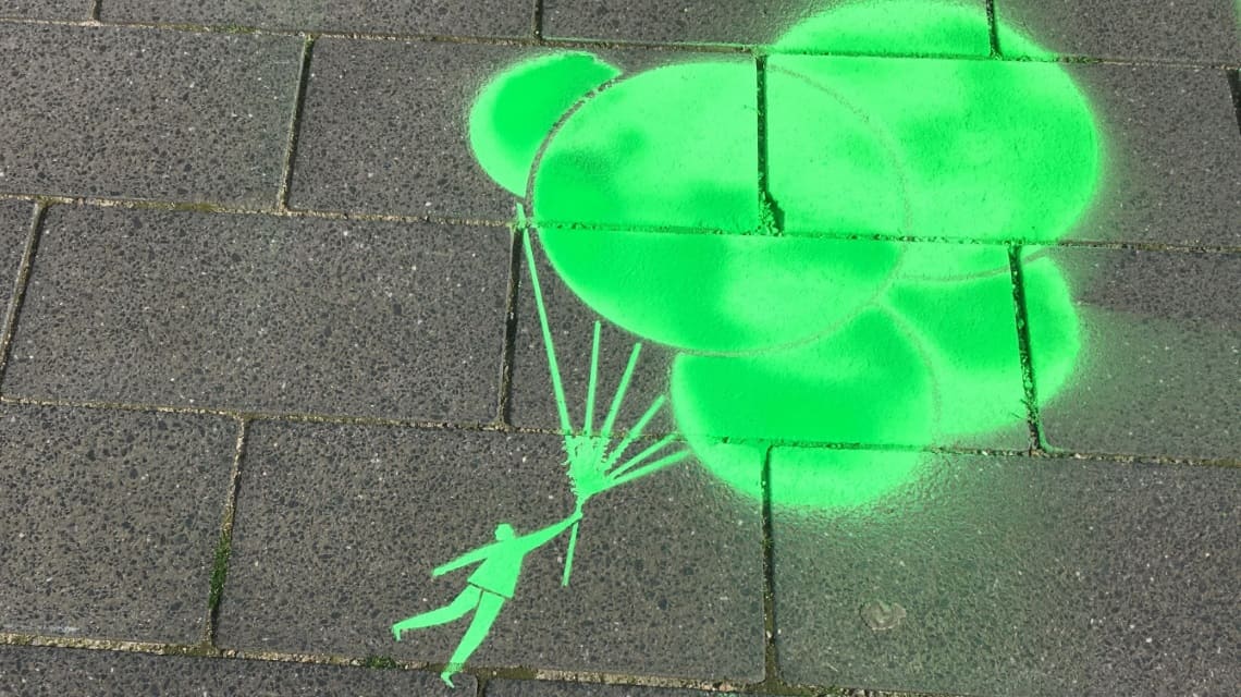 Mit Kreide in knallgrün auf Asphalt gesprühte Grafik: Ein Mensch hängt an einer Traube Luftballons und schwebt 