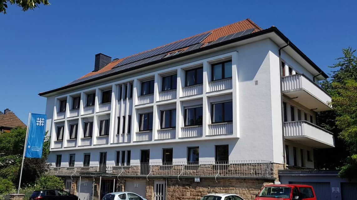 Das zentrale Büro, ein dreistöckiges, weißes Gebäude mit Solarplatten auf dem Dach vor blauem Himmel. Davor einige Autos und ein Fahnenmast mit blauer Kirchentagsfahne