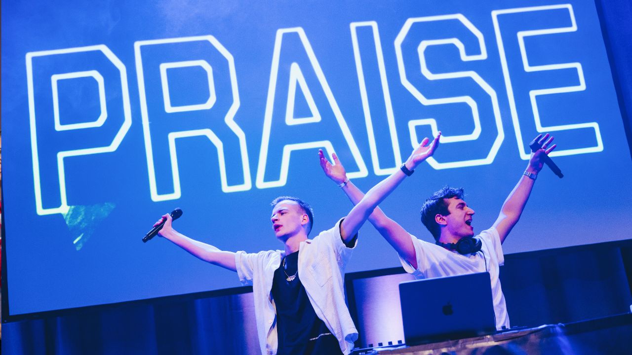 Zwei junge Menschen stehen mit Mikrofonen und DJ-Pult auf einer Bühne. Im Hintergrund ist groß das Wort „Praise“ zu sehen.
