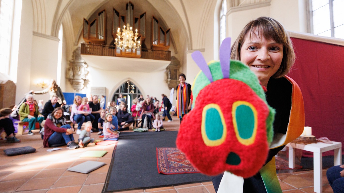 Eine Frau hält die Raupe Nimmersatt aus Stoff in die Kamera, während im Hintergrund eine Gruppe von Menschen in einer Kirche sitzt.