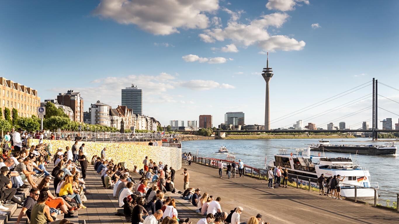 Menschen sitzen auf der Freitreppe am Rheinufer in Düsseldorf. Im Hintergrund sind der Rhein, der Rheinturm und die Rheinkniebrücke zu sehen.