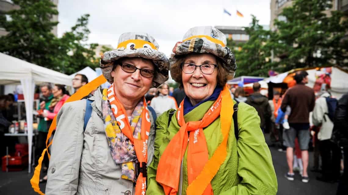 Zwei Damen mit bunten Hüten aus Zeitungen und orangen Schals lachen in die Kamera beim Kirchentag in Berlin 2017 (Abend der Begegnung)