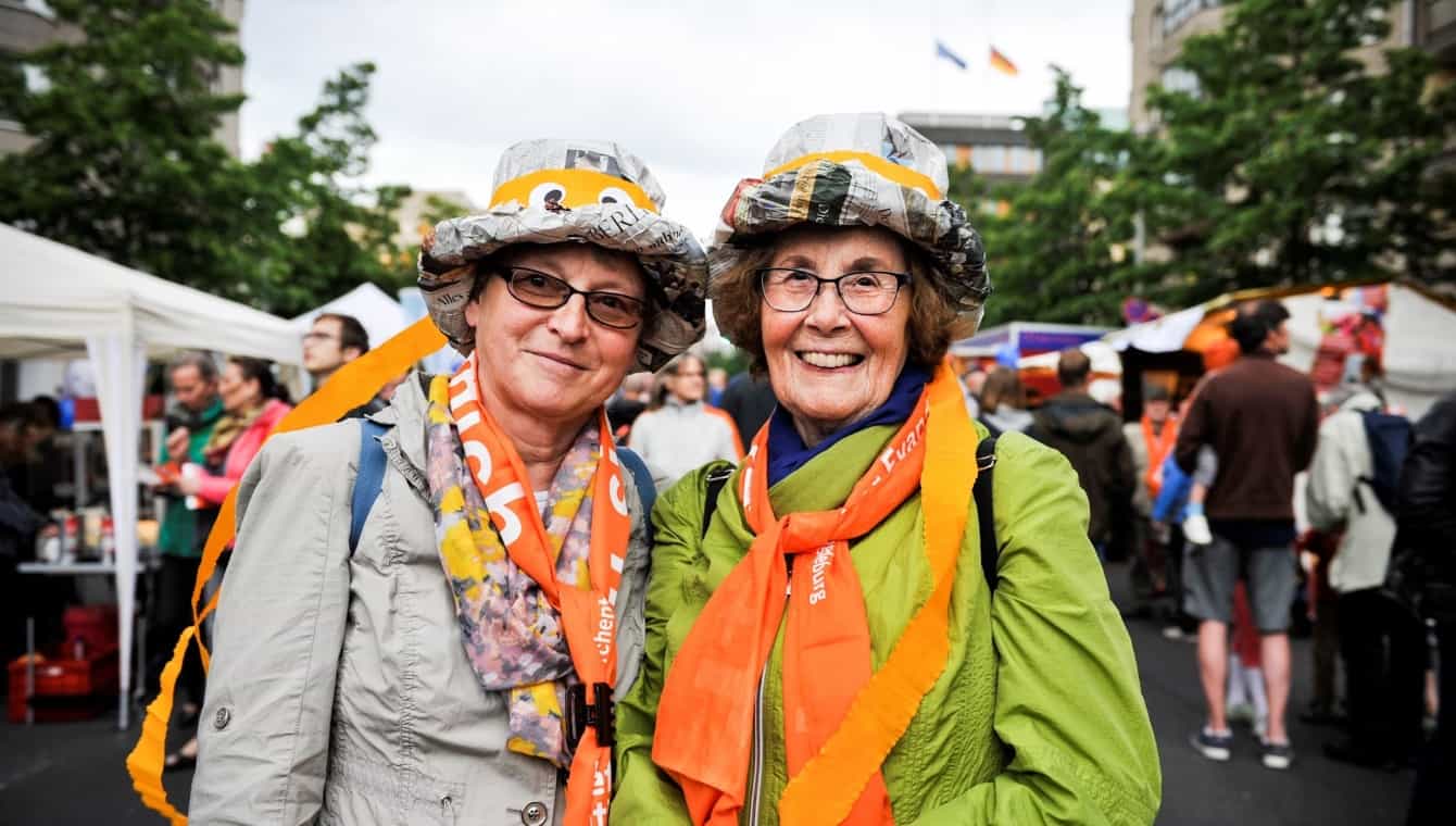 Zwei Damen mit bunten Hüten aus Zeitungen und orangen Schals lachen in die Kamera beim Kirchentag in Berlin 2017 (Abend der Begegnung)