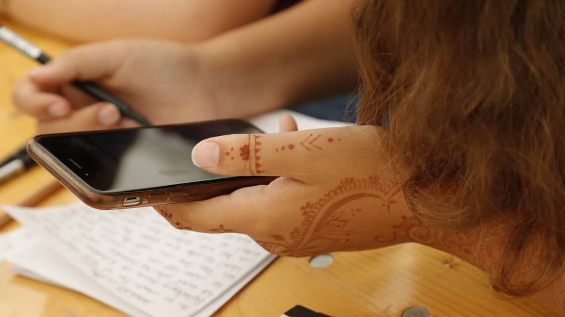 Die mit Henna bemalte Hand eines Mädchens mit einem Smartphone, im Hintergrund ihre andere Hand mit Zettel und Stift 
