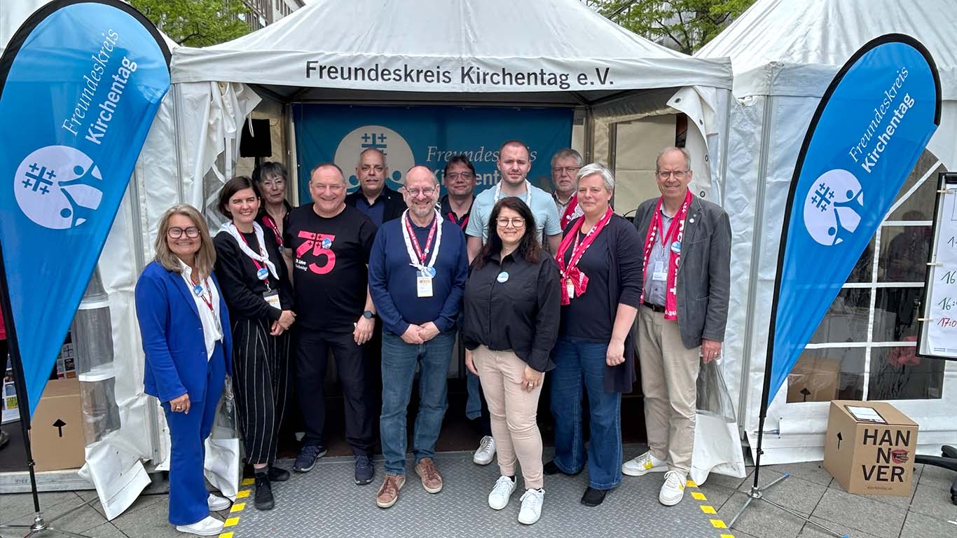 Zwölf Menschen verschiedenen Alters posieren an einem Stand mit Kirchentagsdekoration