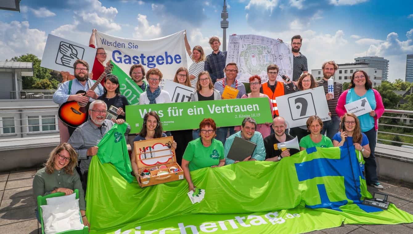Mitarbeitende Dortmund (Foto: DEKT/Stephan Schütze) Gruppenfoto auf einer Dachterrasse. Die Mitarbeitende halten viele unterschiedliche Gegenstände (Gitarre, Moderationskoffer, Schild "Was für ein Vertrauen, Stadtplan, Schilder mit Piktogrammen, Aktenordner, Megaphon...). Im Hintergrund der Dortmunder Fernsehturm