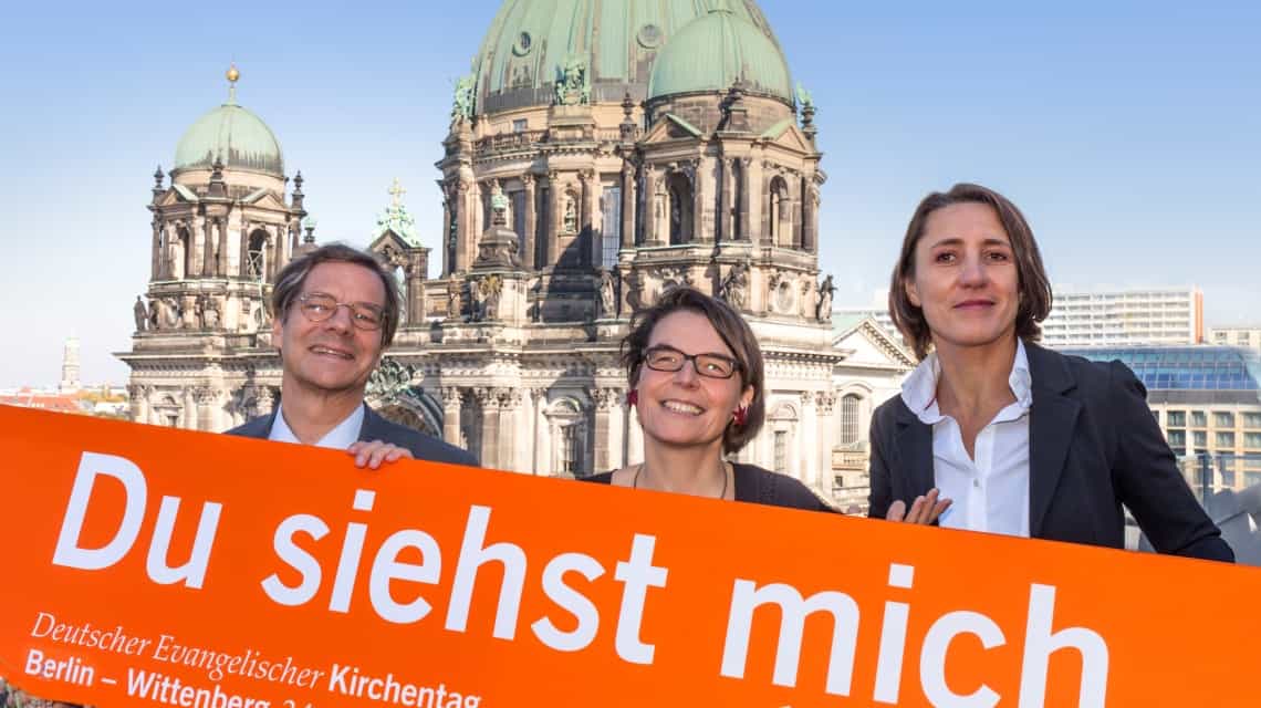 Christina Aus der Au, Ellen Ueberschär, Markus Droege (links) vor dem Berliner Dom mit dem einem großen Schild auf dem "Du siehst mich" in weiß auf orange steht