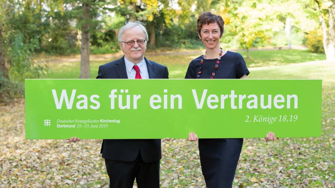 Hans Leyendecker und Julia Helmke stehen in einem Park und halten ein großes Schild mit der Losung in weiß auf grün "Was für ein Vertrauen"