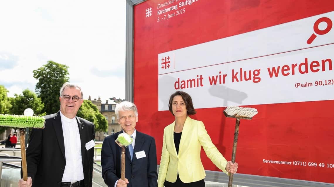 Frank Otfried July, Andreas Barner und Ellen Ueberschär vor einer großen Plakatwand mit dem Motiv "damit wir klug werden". Sie haben Schrubber in der Hand, weil sie plakatiert haben