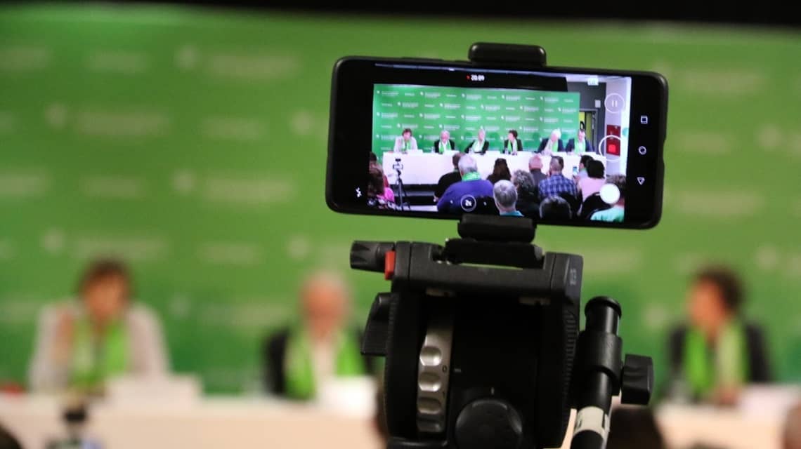 Ein Smartphone auf einem Stativ filmt das Podium einer Pressekonferenz beim Kirchentag in Dortmund. 6 Personen sind verschwommen vor der grünen Pressewand zu sehen