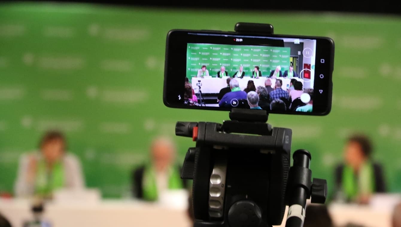 Ein Smartphone auf einem Stativ filmt das Podium einer Pressekonferenz beim Kirchentag in Dortmund. 6 Personen sind verschwommen vor der grünen Pressewand zu sehen