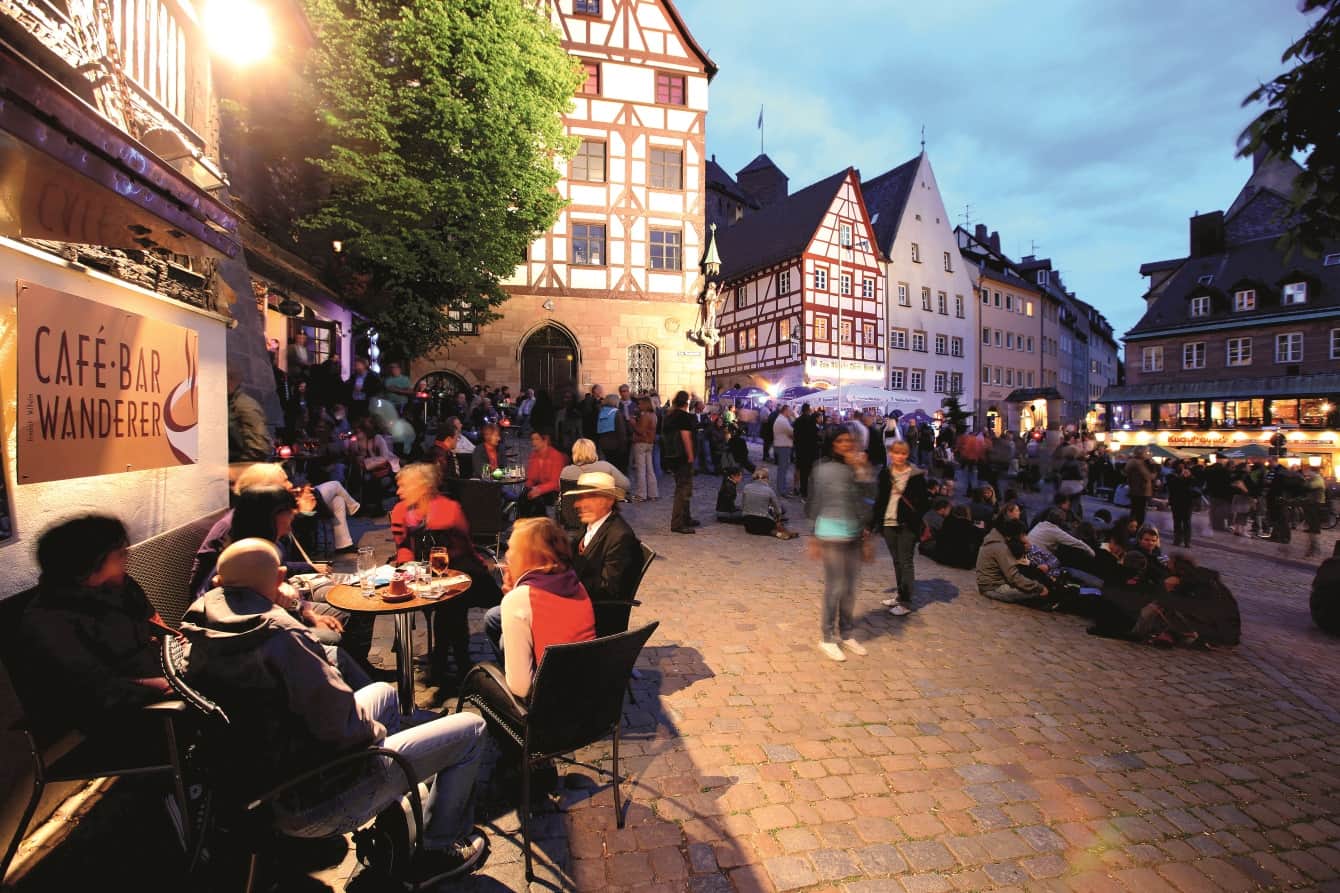 Offener Platz zwischen angestrahlten Fachwerkhäusern, viele Menschen sitzen an Tischen, auf dem Boden oder stehen zusammen