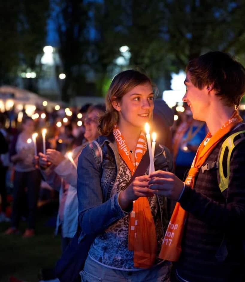 Ein junges Paar schaut sich lächelnd in die Augen während sie die weißen Kerzen zum Abendsegen angezündet zwischen sich halten