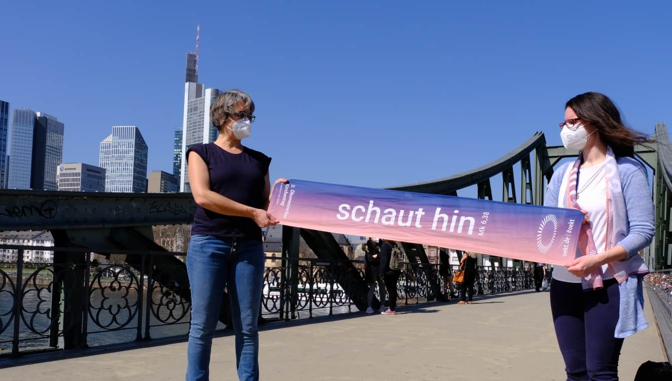 Zwei Frauen mit FFP2 Maske halten Abstand indem sie zwischen sich den "schaut hin" Schal halten, sie stehen auf einer Brücke über den Main, im Hintergrund die Frankfurter Skyline