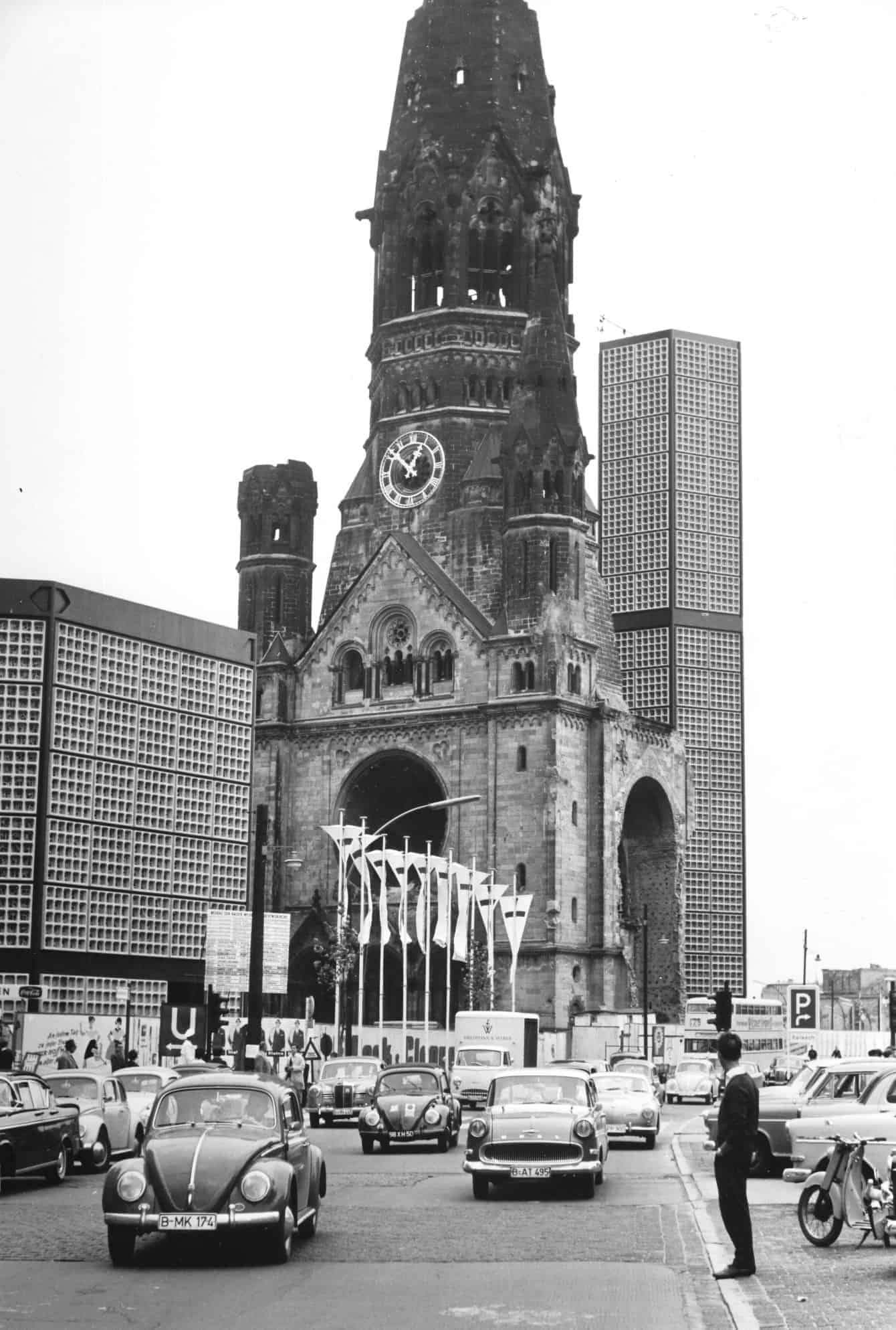 Schwarz-weiß Aufnahme der zerstörten Kaiser-Wilhelm-Gedächtniskirche, im Hintergrund der moderne Neubau, vor der Kirche viele Kirchentagsfahnen und Autos, z.B. VW Käfer