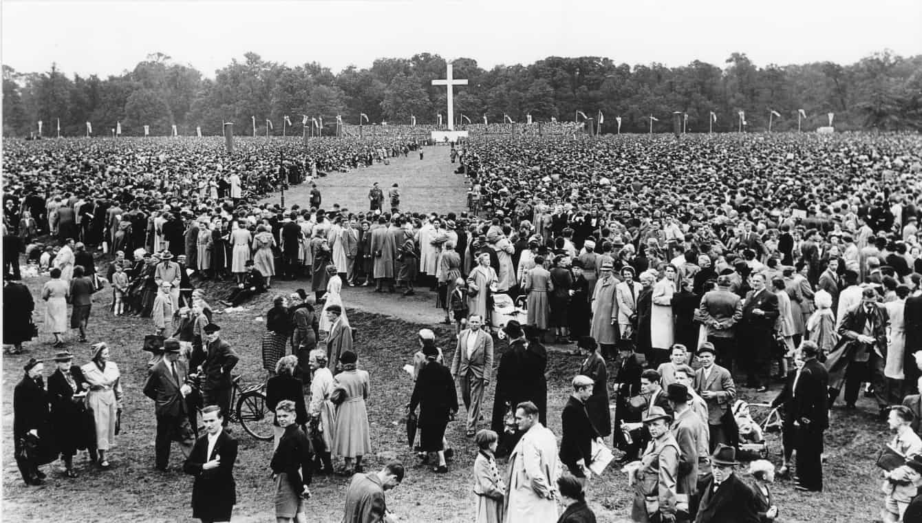 Schwarz-weiß Aufnahme einer sehr großen Menschenmenge, Schlussgottesdienst beim Kirchentag in Leipzig 1954 - mit 650.000 Besucher*innen die bis heute größte protestantische Versammlung in Deutschland, mittig eine Schneise zu einem großen weißen Kreuz, vorne einzelne Personen in kleinen Gruppen, einer ist mit dem Fahrrad da