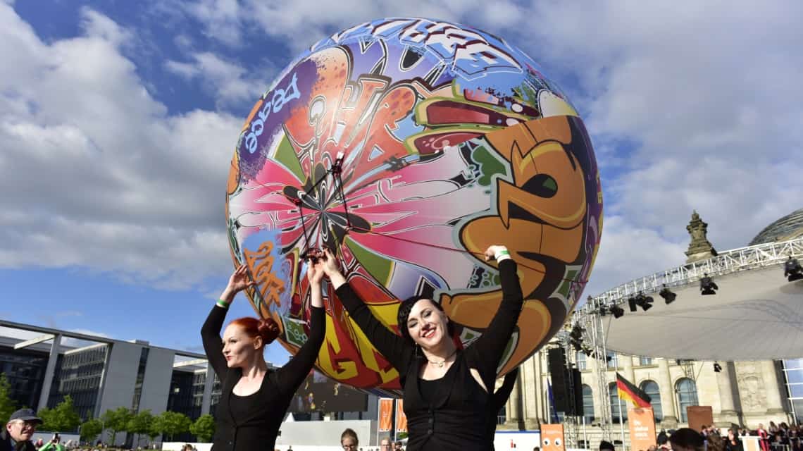 Performance mit einem riesigen Ball mit bunten Graffitis bedruckt, drei schwarz gekleidete Frauen recken ihn in die Höhe, eine lächelt direkt in die Kamera