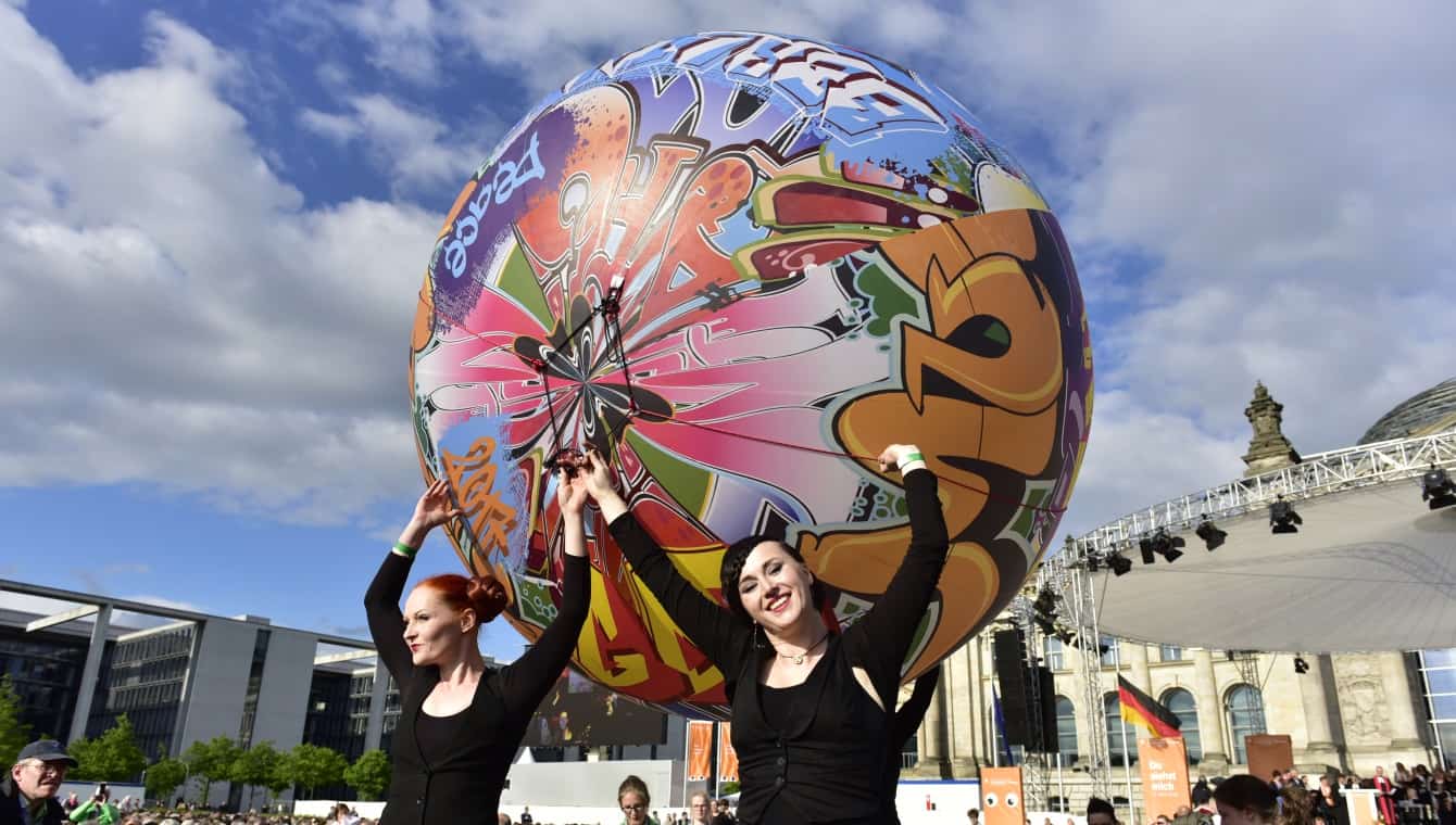 Performance mit einem riesigen Ball mit bunten Graffitis bedruckt, drei schwarz gekleidete Frauen recken ihn in die Höhe, eine lächelt direkt in die Kamera