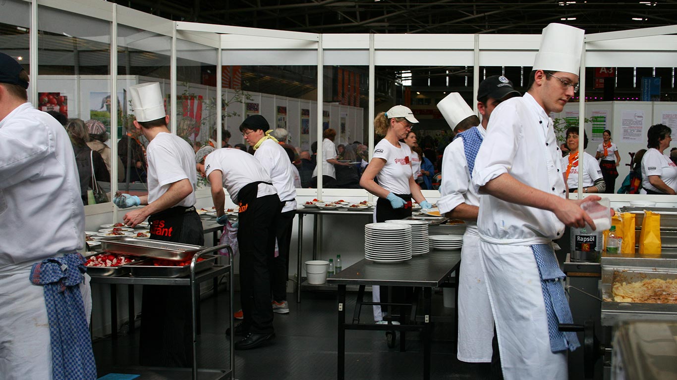 Blick in die Küche. Köch:innen in weißen Schürzen und Kochmützen bei unterschiedlichen Aufgaben.
