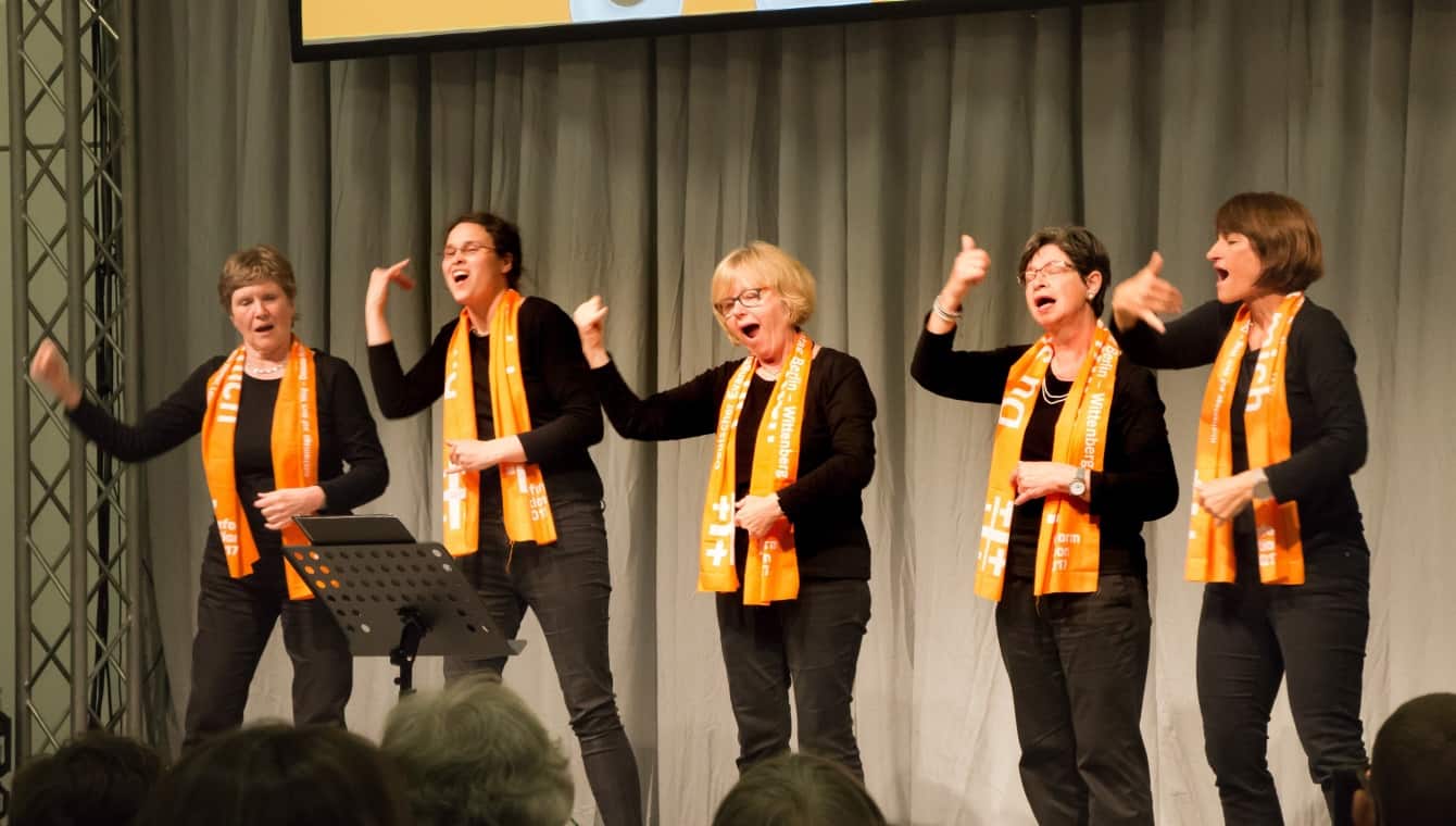 Fünf Frauen singen auf der Bühne in Gebärden, sie sind ganz in schwarz gekleidet und tragen den orangenen Schal des Kirchentages
