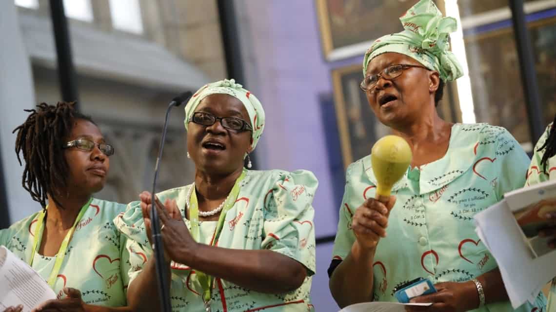 Drei internationale Mitwirkende bei ihrem Auftritt beim Kirchentag, vermutlich afrikanischer Gospel, sie tragen hellgrüne Gewänder, zwei haben passende Kopftücher, eine Rastazöpfe. Die Dame in de Mitte klatscht im Takt, die rechts spielt Maracas