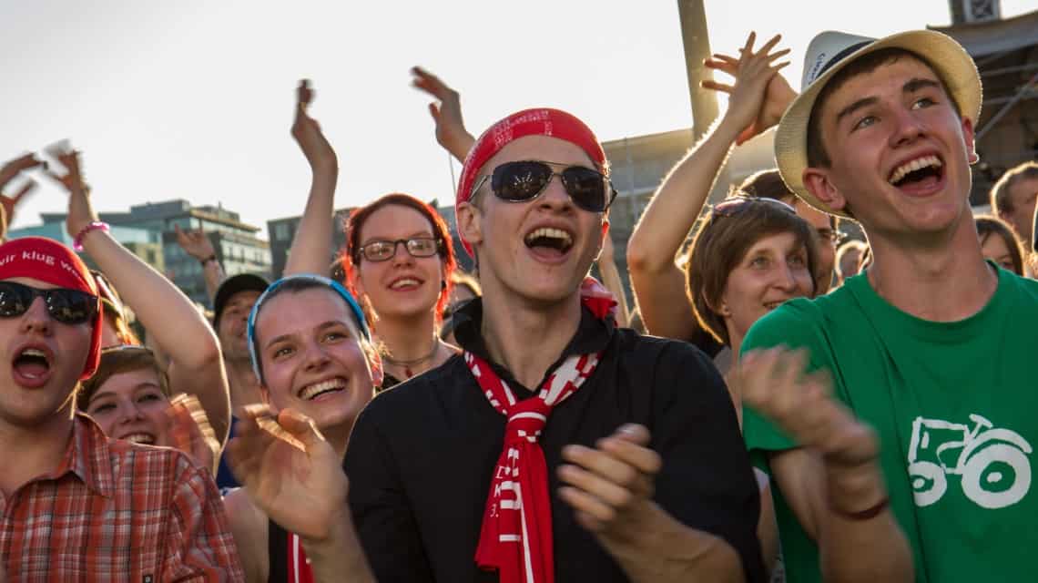 Jugendliche Konzertbesucherinnen beim Kirchentag in Stuttgart, sie lachen, rufen und klatschen ausgelassen, der Junge in der Mitte wirkt besonders begeistert: er trägt den roten Schal und das rote MuFuTu auf dem Kopf und eine Sonnenbrille