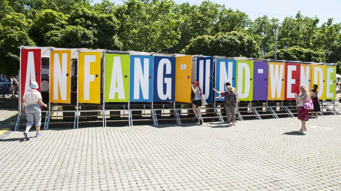 Komposttoiletten, die Türen sind farbig und darauf steht ein großer Buchstabe, so dass sich zusammen ein Regenbogen mit dem Spruch "Anfang und Wende" ergibt